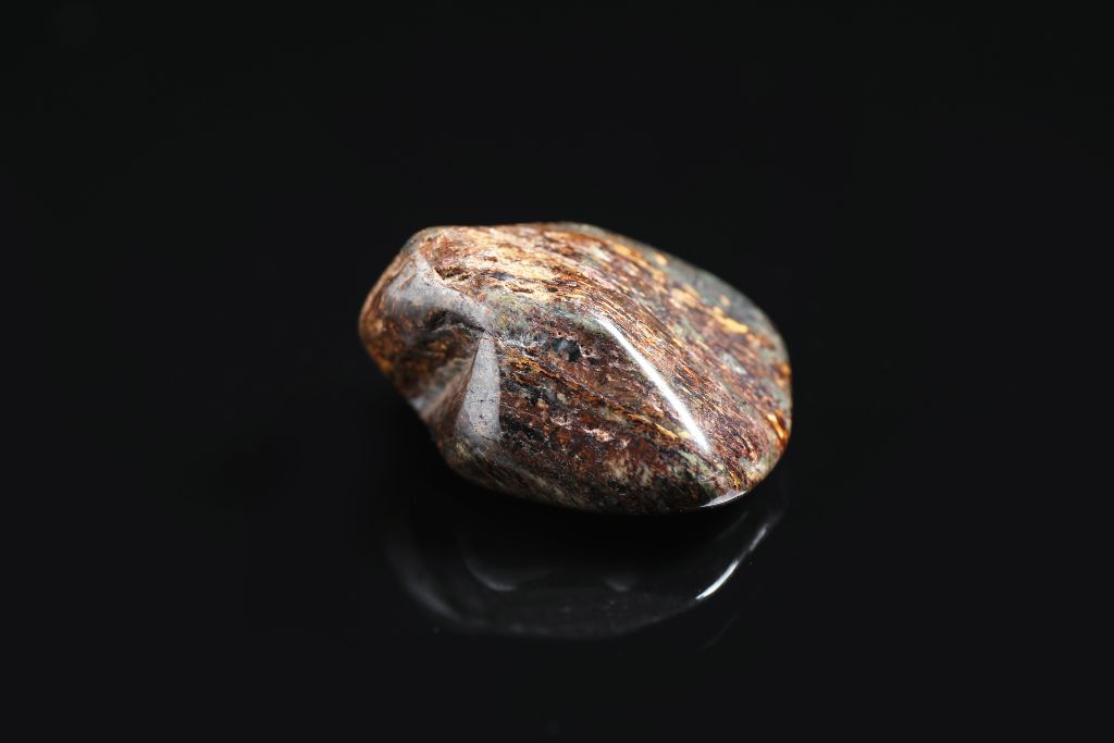 The image displays a single polished stone with earthy brown, reddish, and dark gray tones, resting on a sleek black surface. The stone's smooth surface reflects a subtle shine, and its marbled pattern features streaks of lighter and darker colors, giving it a rich, organic appearance.