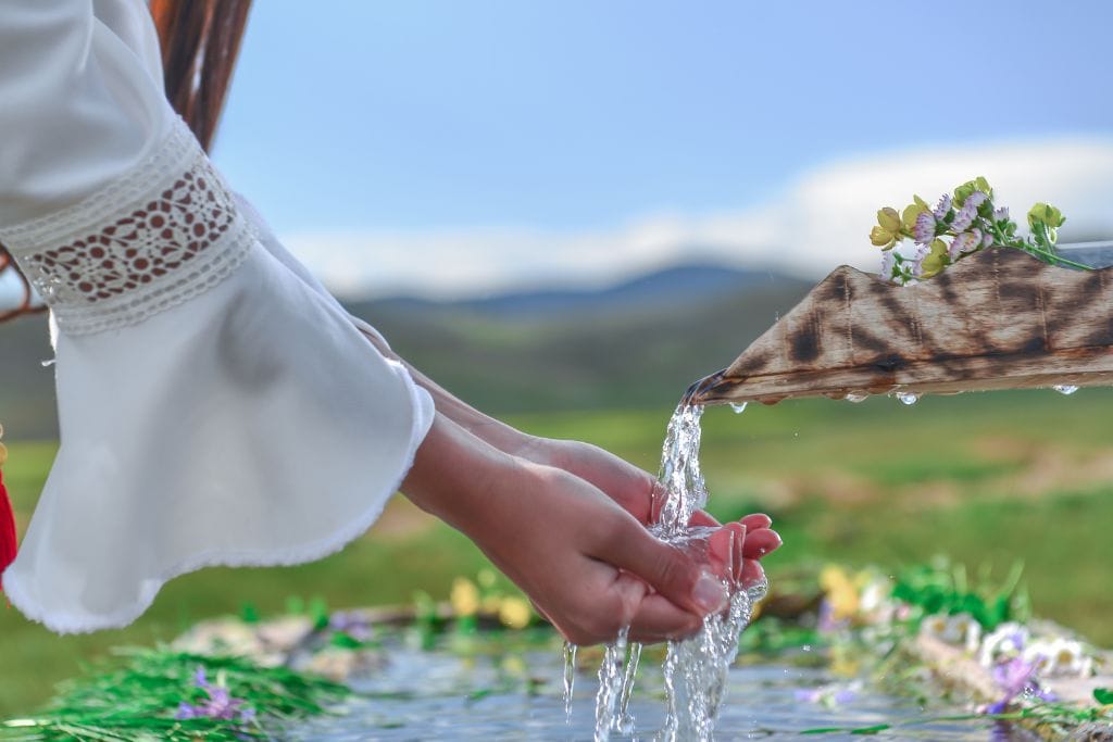 The image shows a person in a white, long-sleeved garment with lace detail, cupping their hands under a stream of water flowing from a wooden spout decorated with small flowers. The scene takes place outdoors, with a blurred green landscape and mountains in the background, evoking a peaceful and natural atmosphere.
