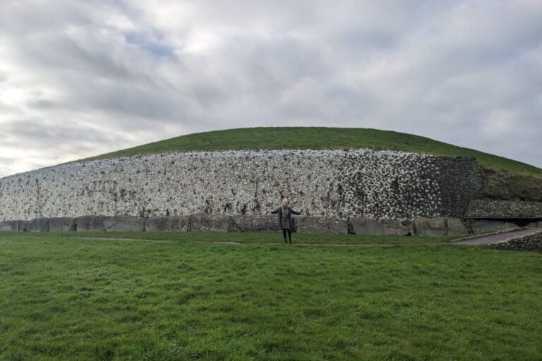 This image shows a large ancient structure, most likely the Newgrange passage tomb in Ireland. The grass-covered mound features a distinctive stone wall made of white quartz and other dark stones, with a person standing in front for scale. The cloudy sky adds to the atmosphere of the site.