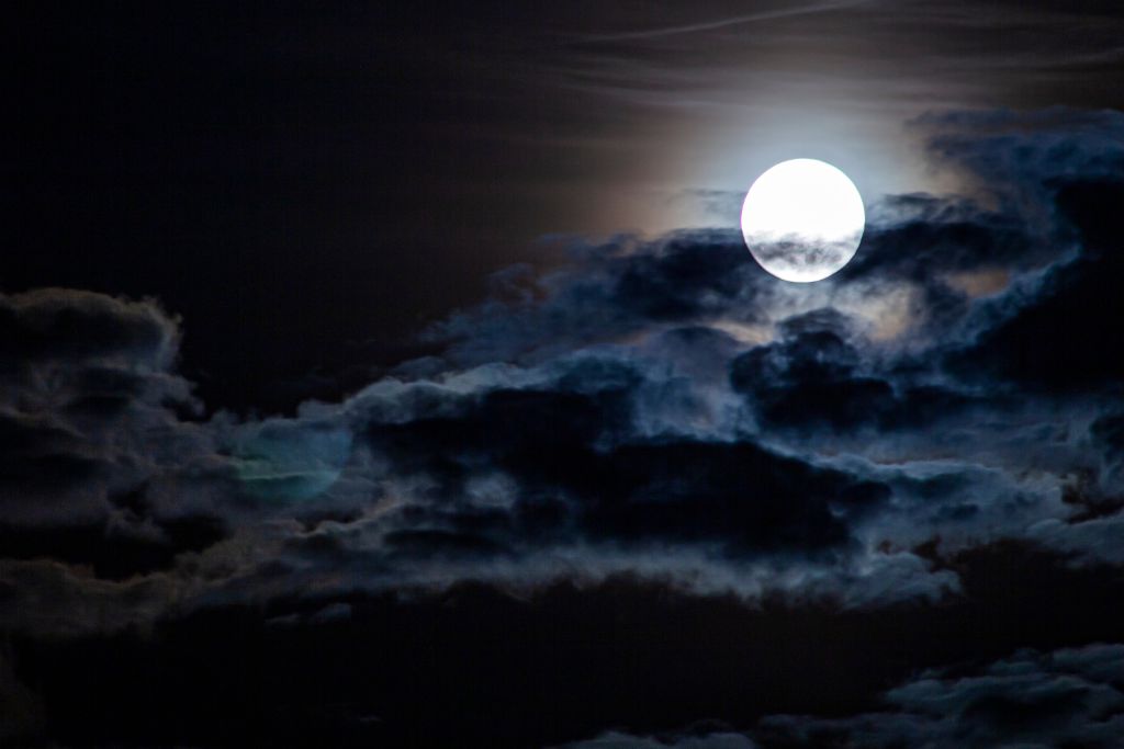 The image captures a full moon shining brightly through a partially cloudy night sky. Dark, thick clouds with soft edges surround the moon, which illuminates the scene with an eerie, ethereal glow. The contrast between the deep blacks and the moon's bright light adds a mysterious, dramatic atmosphere.