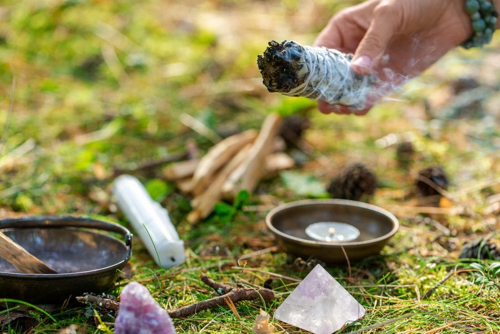 The image shows a person holding a smoldering bundle of sage, releasing smoke as part of a ritual or cleansing ceremony in an outdoor forest setting. Nearby, various items are arranged on the ground, including a burning candle in a dish, a small pyramid-shaped crystal, a white candle, and wooden sticks. The background features soft greenery and pinecones, adding to the natural, peaceful ambiance.