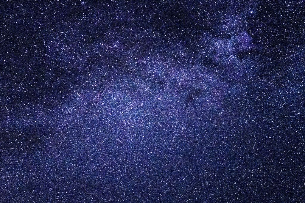 The image shows a breathtaking view of a star-filled night sky, possibly capturing the Milky Way galaxy. Countless stars twinkle across the vast expanse, creating a deep blue and purple cosmic tapestry. The density of stars and faint nebulae give the sky a textured, almost ethereal appearance, evoking a sense of wonder and infinity.