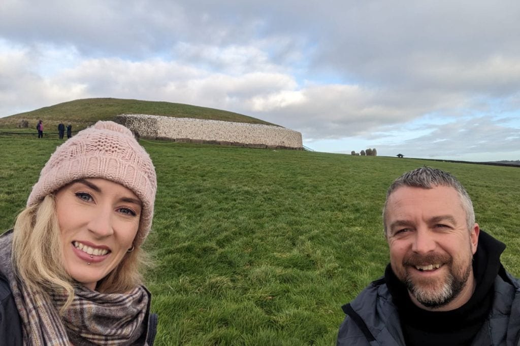 This image is a selfie of two people smiling in front of the Newgrange passage tomb in Ireland. The ancient mound is visible in the background, covered in grass with its distinct stone wall partially visible. The sky is partly cloudy, and other visitors can be seen in the distance exploring the site.