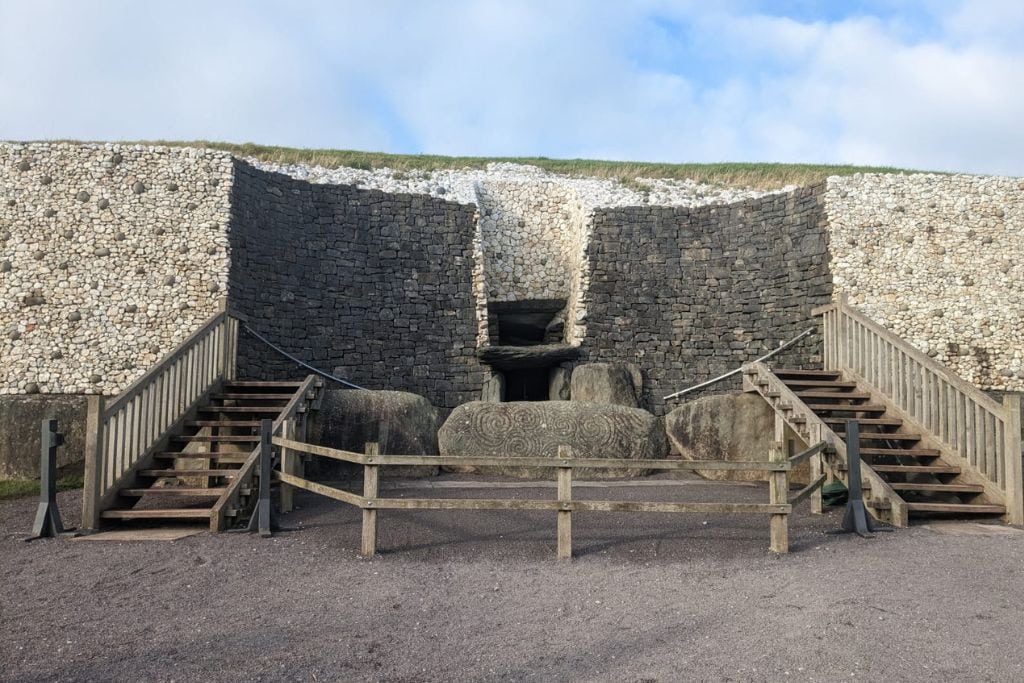 This image shows the entrance to the Newgrange passage tomb in Ireland, featuring two wooden staircases leading up to the stone entrance. The iconic entrance stone, engraved with Neolithic spiral patterns, is positioned at the base. The structure's outer wall consists of light-colored quartz and dark stones, with the mound rising above. The sky is partly cloudy, adding depth to the scene.