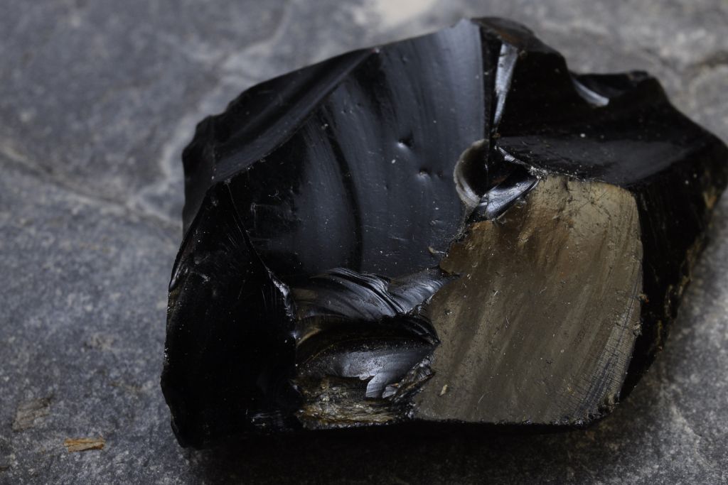 The image displays a close-up of a rough, glossy black stone, likely obsidian, resting on a gray surface. The stone has sharp, jagged edges, and its highly reflective surface shows both smooth and fractured textures. One side of the stone reveals a beige, grainy interior where it appears to have been chipped or broken.