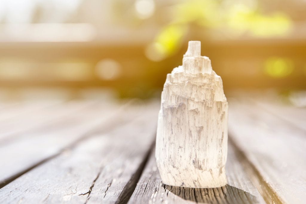 The image shows a tall, cylindrical selenite crystal standing upright on a wooden surface. The crystal's translucent white structure appears layered and rough, with a pointed top. The background is blurred, with warm, golden light filtering in, creating a soft and serene atmosphere.