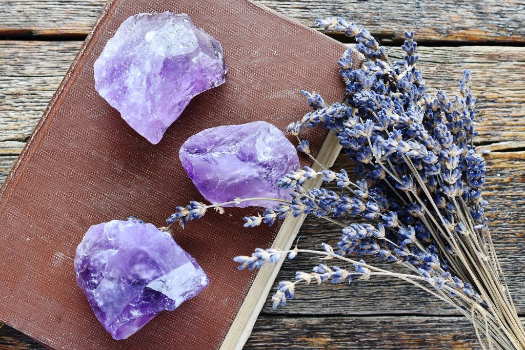 The image shows three rough amethyst crystals placed on top of a closed brown book, alongside a dried bundle of lavender flowers. The scene is set against a rustic wooden surface, giving a natural, earthy vibe. The purple hue of the amethysts complements the soft blue tones of the lavender, creating a visually calming and harmonious composition.