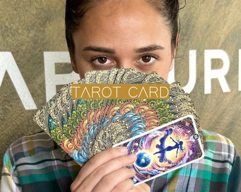 The image shows a person holding up a fan of intricately designed tarot cards, with one card featuring a cosmic, mystical illustration visible. The text "TAROT CARD" is overlaid in bold, stylized letters on a gold rectangle in the center of the image.