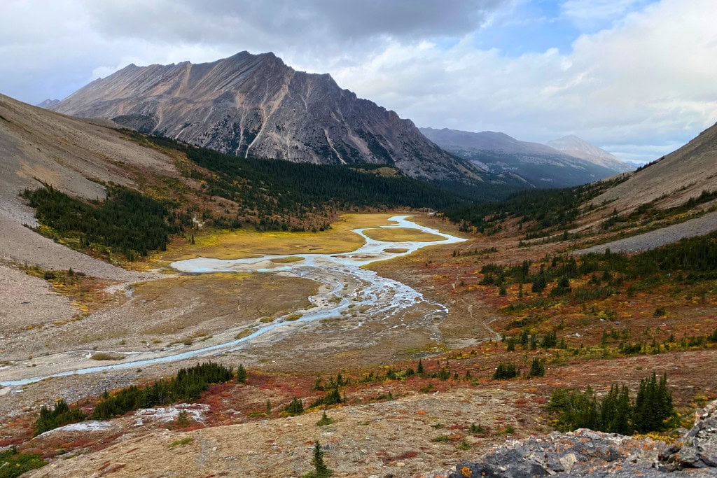 Is Banff Worth Visiting? Explore This Spiritual Place in 2026 A scenic valley view during a multi-day hike on the border of Banff and Jasper National Parks. A winding river flows through the vibrant landscape of autumn foliage, surrounded by rocky mountains and forested slopes under a cloudy sky.