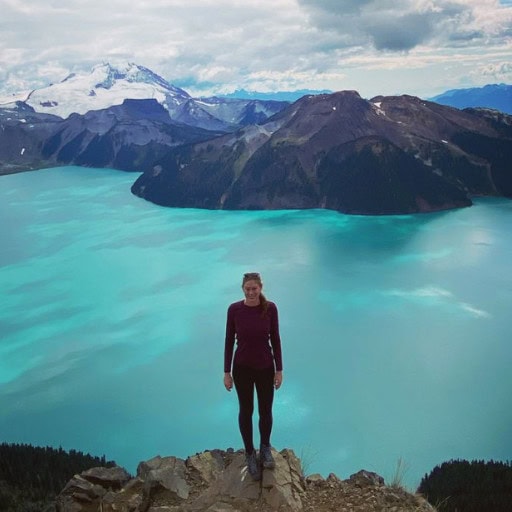 Is Banff Worth Visiting? Explore This Spiritual Place in 2026 A hiker standing at the edge of a cliff overlooking a stunning turquoise lake and mountainous landscape in Banff National Park. Snow-capped peaks rise in the background under a cloudy sky, showcasing the breathtaking beauty of Banff.
