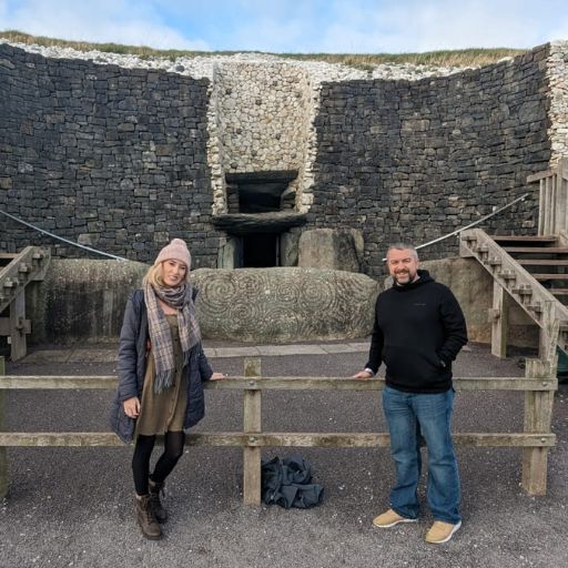 This image shows two people standing in front of the entrance to the Newgrange passage tomb in Ireland. They are positioned next to the iconic entrance stone, engraved with ancient spiral designs. Behind them are two wooden staircases that lead to the passageway, with the stone walls of the tomb visible in the background. The individuals are smiling, enjoying their visit to this historic site.