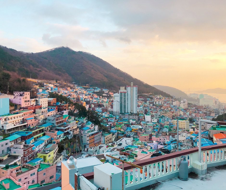 Panoramic view of colorful houses and tall buildings nestled against a hillside in Busan at sunset, showcasing the vibrant charm of the city. The scenic landscape highlights why Busan is worth visiting for its unique blend of urban life and nature.