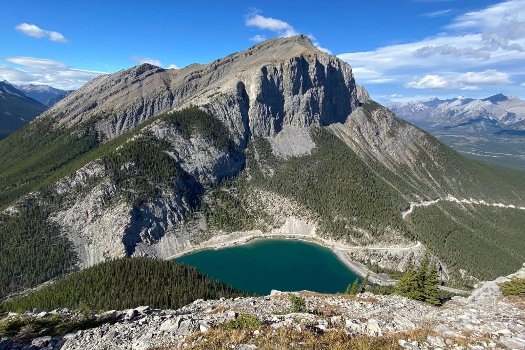 Is Banff Worth Visiting? Explore This Spiritual Place in 2026 A stunning view of Mount Rundle and a deep blue lake as seen from the Ha Ling Peak hike near Banff. The rugged mountain rises dramatically above the forested landscape, with clear skies and distant mountain ranges in the background.