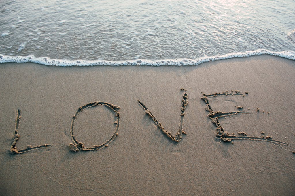 Angel Number 0000: Spiritual Messages & Symbolic Meaning A heart symbol drawn in the wet sand on a seashore, with the word 'Love' written inside the heart. Gentle waves approach the shore, with soft brown sand beneath and a calming, romantic atmosphere.