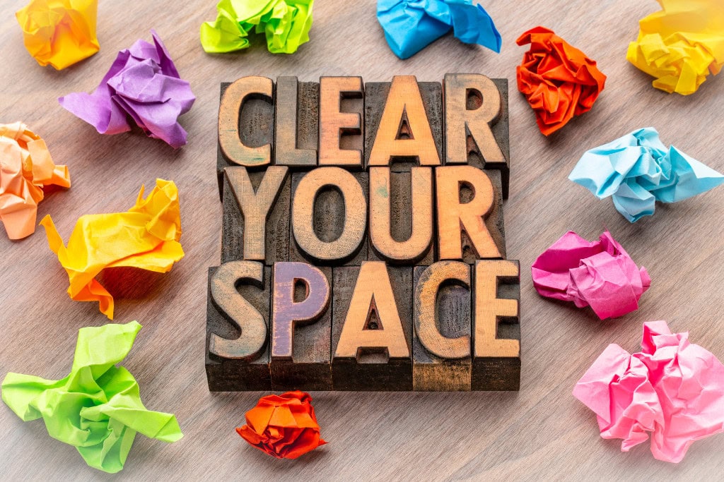 Cleansing Your House from Negative Energy: Everything You Need to Know! This image features the words "CLEAR YOUR SPACE" in bold, wooden block letters set on a wooden surface. Surrounding the text are crumpled pieces of brightly colored paper in shades of purple, blue, yellow, orange, and green, suggesting the idea of decluttering or organizing. The overall feel is playful and motivational.