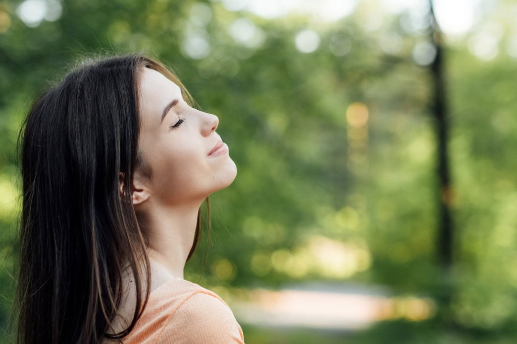 Cleansing Your House from Negative Energy: Everything You Need to Know! This image shows a young woman with her eyes closed, tilting her head slightly upwards, as if enjoying a moment of peace or deep breath in nature. The soft, blurred green background suggests she is outdoors, surrounded by trees or in a park. The overall mood is one of relaxation, mindfulness, and connection with nature.