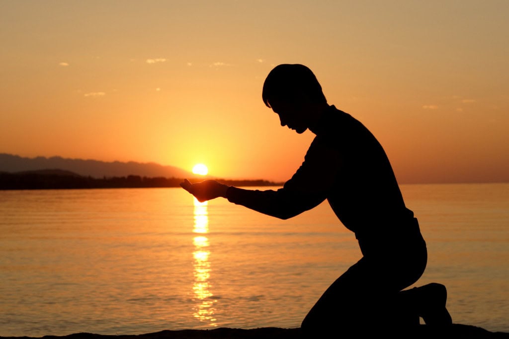 Cleansing Your House from Negative Energy: Everything You Need to Know! This image features a silhouetted person kneeling by the water at sunset, holding their hands up as if cradling the sun. The golden light reflects off the water, creating a peaceful and contemplative scene. The image evokes a sense of connection with nature, mindfulness, or perhaps a symbolic gesture of holding the sun's energy.
