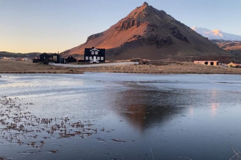 A scenic view of a small black house with white trim near a frozen lake, reflecting the surrounding landscape. Behind the house, the sun casts a warm glow on a rugged mountain, with the snow-capped Glacier Snaefellsjokull visible in the distant background, adding contrast to the rocky terrain. The image captures the peaceful atmosphere of Iceland’s natural beauty.