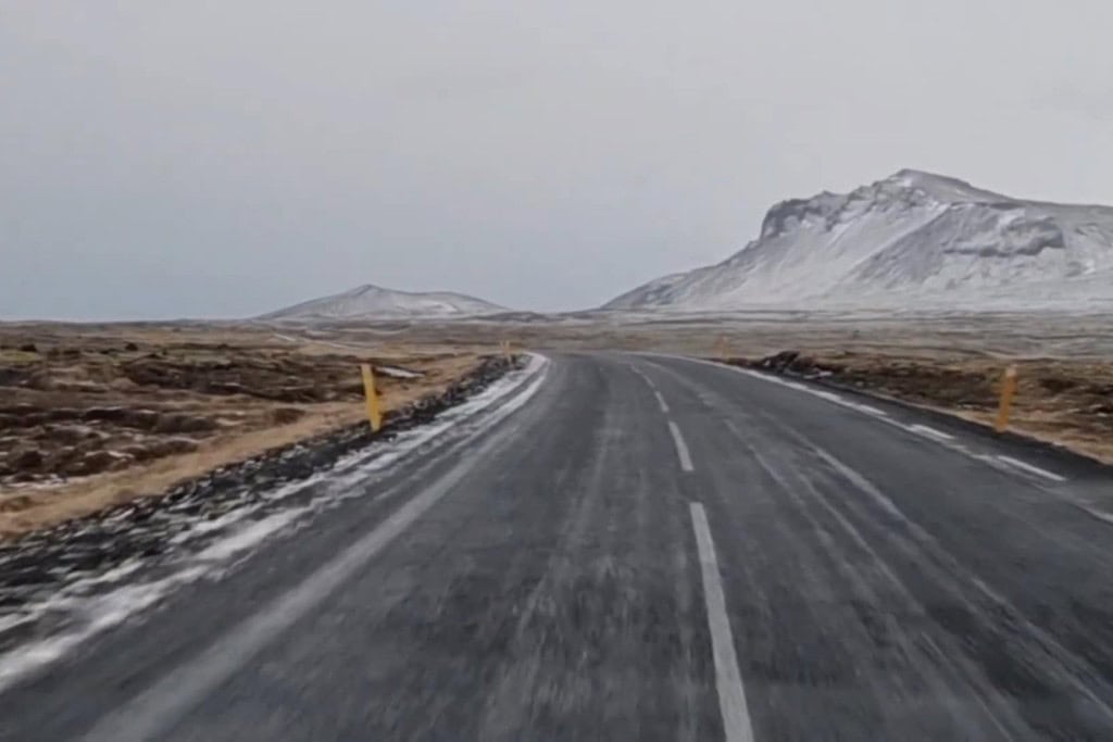 Glacier Snaefellsjokull: Your Guide to Center of Earth! A solitary road stretches forward through a vast, barren landscape dusted with snow, leading towards rugged, snow-capped mountains under a pale, overcast sky. Yellow markers line the roadside, enhancing the feeling of isolation in this stark, icy terrain. The scene captures the remote, tranquil beauty of Iceland’s winter roads.