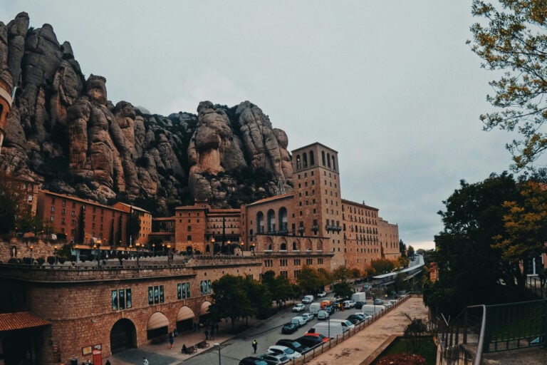 A panoramic view of the Montserrat Monastery nestled against the backdrop of jagged rocky mountains. The large, historic stone buildings contrast with the natural cliff formations, with cars and visitors scattered around the monastery's grounds, creating a lively atmosphere.