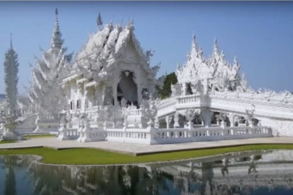 This image shows the stunning Wat Rong Khun, also known as the White Temple, located in Chiang Rai, Thailand. The intricately designed white structure is reflected in a nearby pond, highlighting its ornate carvings and statues. The bright, clear sky provides a striking backdrop to the temple's delicate and elaborate details.