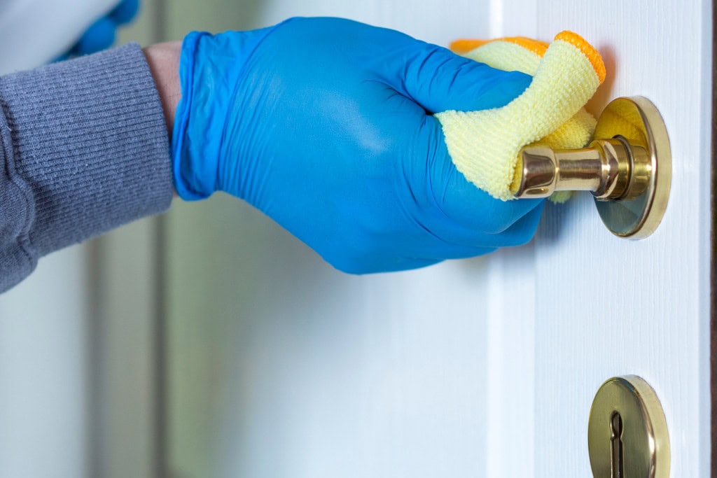 Cleansing Your House from Negative Energy: Everything You Need to Know! This image shows a person wearing blue gloves cleaning a brass door handle with a yellow microfiber cloth. The action suggests disinfecting or sanitizing, with a focus on hygiene and cleanliness. The door handle and lock are polished, and the overall composition conveys a sense of careful attention to cleaning.