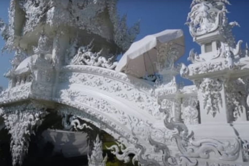 Wat Rong Khun: The White Temple Of Chiang Rai, Thailand This close-up image showcases the intricate architectural details of Wat Rong Khun (the White Temple) in Chiang Rai, Thailand. The white, ornate carvings on the bridge and surrounding structures are elaborate and highly detailed, featuring flowing patterns and mythical figures. The craftsmanship is accentuated by the bright blue sky, which contrasts beautifully with the temple's pristine white surfaces.