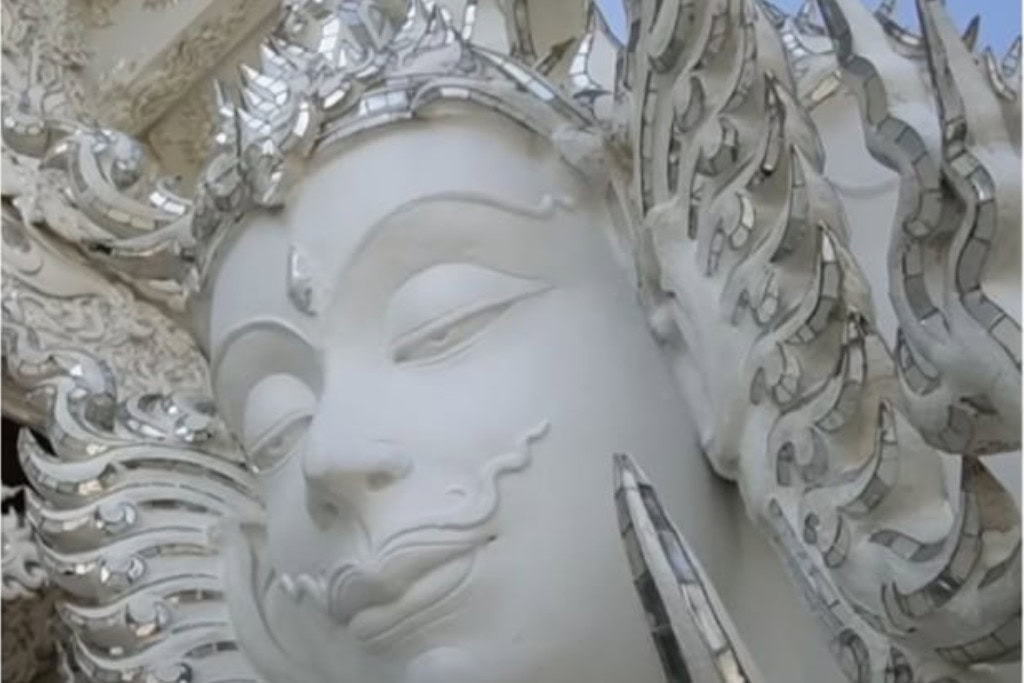 Wat Rong Khun: The White Temple Of Chiang Rai, Thailand This close-up image captures the serene face of a deity statue from Wat Rong Khun (the White Temple) in Chiang Rai, Thailand. The statue is detailed with smooth white surfaces and mirrored elements that reflect light, creating a radiant and peaceful expression. The intricate carvings surrounding the face emphasize the divine and delicate craftsmanship of the temple's design.