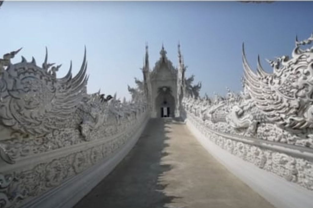Wat Rong Khun: The White Temple Of Chiang Rai, Thailand This image shows a path leading to the main entrance of Wat Rong Khun (the White Temple) in Chiang Rai, Thailand. The walkway is bordered by ornate, sculpted railings adorned with mythical creatures and intricate carvings, drawing the eye toward the temple’s grand façade in the distance. The bright white structure contrasts against the clear blue sky, enhancing the temple's ethereal and otherworldly appearance.