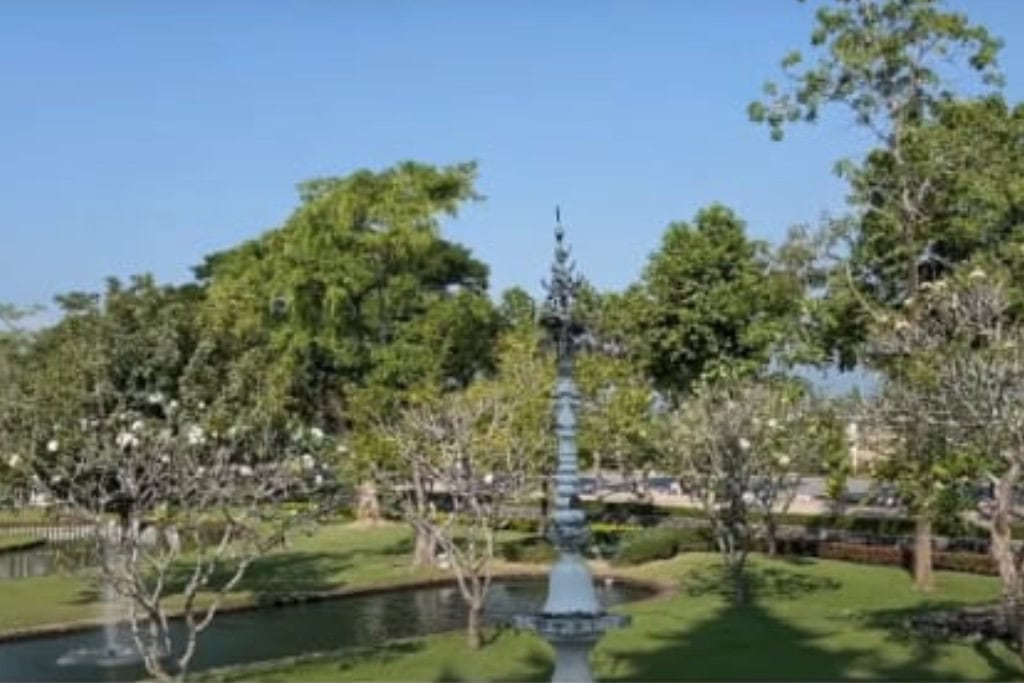 Wat Rong Khun: The White Temple Of Chiang Rai, Thailand This image depicts a peaceful garden scene at Wat Rong Khun (the White Temple) in Chiang Rai, Thailand. The lush greenery, including trees and blooming white flowers, surrounds a small pond with a decorative fountain. The clear blue sky enhances the serene atmosphere of this landscaped area, offering a tranquil contrast to the temple's ornate and intricate structures.