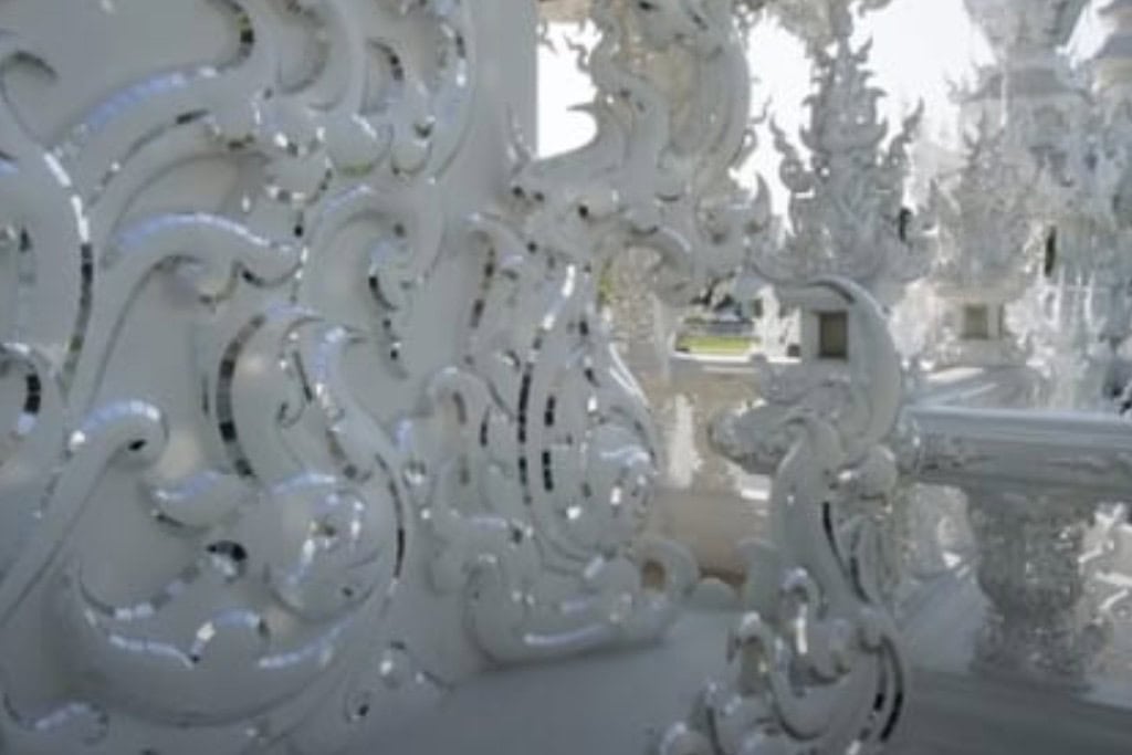 Wat Rong Khun: The White Temple Of Chiang Rai, Thailand This close-up image highlights the intricate decorative patterns on a wall at Wat Rong Khun (the White Temple) in Chiang Rai, Thailand. The swirling, delicate designs are adorned with mirrored glass pieces, which reflect light and enhance the temple's ethereal appearance. The detailed craftsmanship exemplifies the temple's blend of traditional and modern Thai art, creating an ornate and luminous effect.