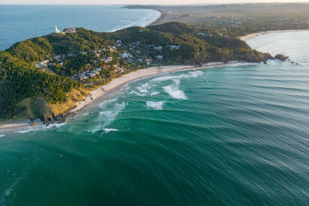 This image appears to be an aerial view of a coastal landscape, showcasing a scenic beach bordered by lush, green hills. A lighthouse sits atop a hill overlooking the coastline, with gentle waves breaking on the shore. The view extends into the horizon, capturing the natural beauty of the area. If this is part of a blog post, feel free to let me know if you'd like specific keywords or further optimization for SEO purposes.