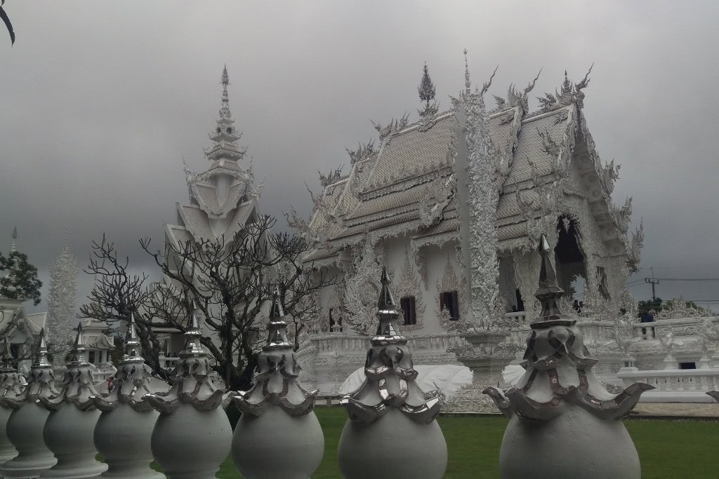 Wat Rong Khun: The White Temple Of Chiang Rai, Thailand This image shows Wat Rong Khun (the White Temple) in Chiang Rai, Thailand, under a cloudy sky. The temple's intricate white structure, adorned with ornate carvings and statues, contrasts with the dark, overcast clouds above. A row of decorative spires lines the foreground, while leafless trees add a stark element to the scene, enhancing the mystical and serene atmosphere of the temple grounds.