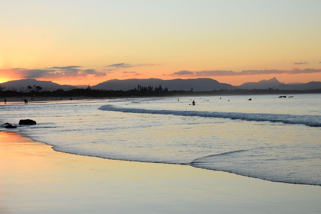 This image features a serene beach at sunset, with soft orange and pink hues filling the sky as the sun sets behind distant mountains. Gentle waves roll onto the shore, and a few people can be seen in the water, likely surfers or swimmers. The peaceful atmosphere is complemented by the silhouettes of beachgoers walking along the shoreline, enjoying the tranquil evening scene. 