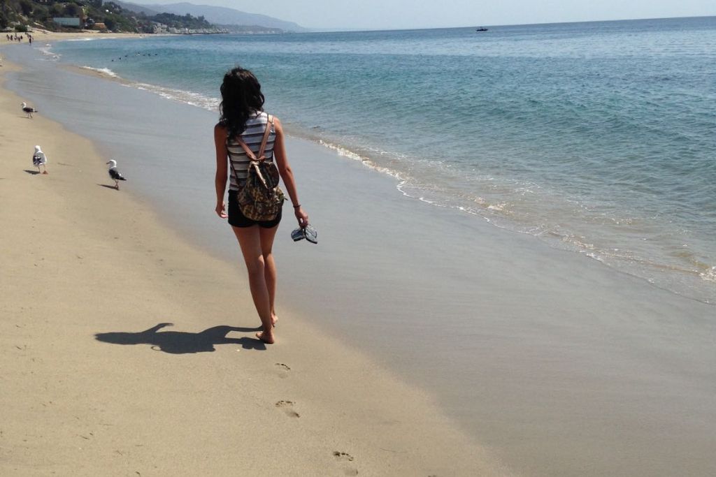 This image captures a woman walking barefoot along a peaceful beach with gentle waves lapping at the shore. She is wearing shorts and a striped tank top, carrying her shoes in one hand, and her backpack rests on her shoulders. Seagulls walk along the sand near her, and the coastline stretches into the distance with calm, blue waters.