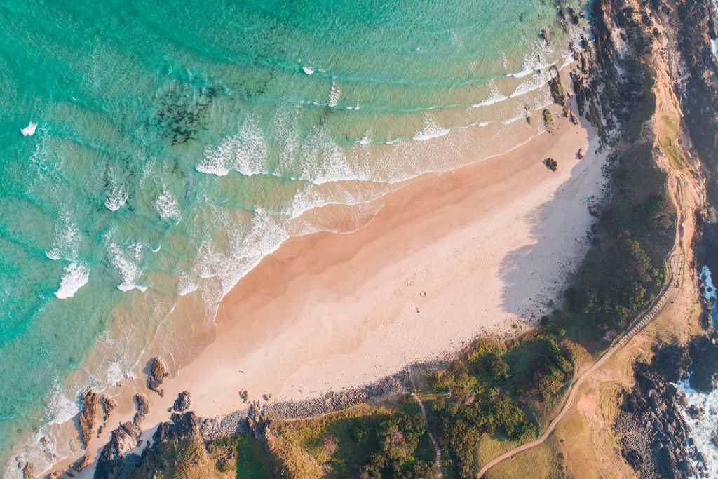 This aerial image shows a stunning stretch of coastline with turquoise waves gently breaking on a sandy beach. The view highlights the contrast between the vibrant ocean, the soft sand, and the rugged cliffs that frame the shore. A walking path winds along the cliffside, offering a scenic route with a view of the untouched beach and ocean below.