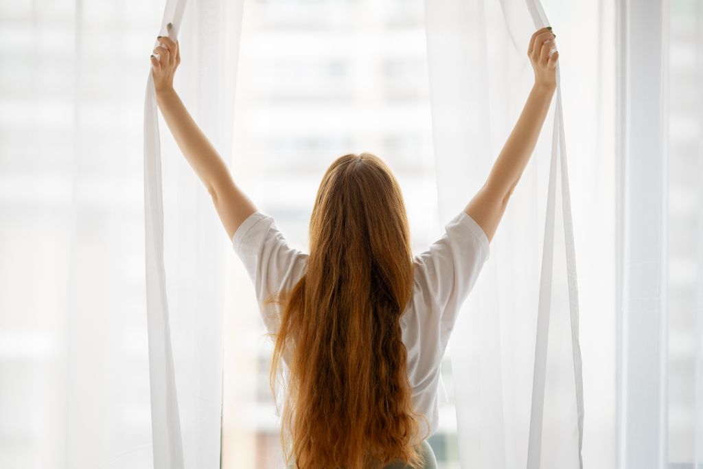 Cleansing Your House from Negative Energy: Everything You Need to Know! This image shows a person with long, wavy hair opening sheer curtains, facing a bright window. The sunlight filters through, creating a peaceful morning atmosphere, with the focus on the person's back and the sense of welcoming a new day.