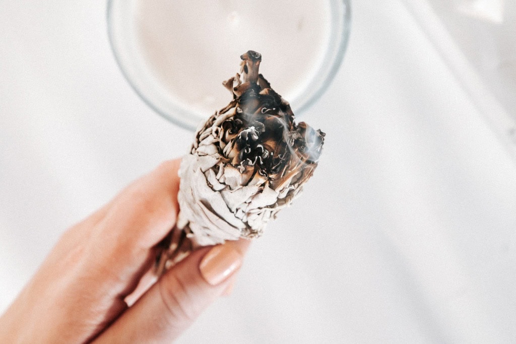 Cleansing Your House from Negative Energy: Everything You Need to Know! This image shows a hand holding a smoldering bundle of sage, with wisps of smoke rising from the burning end. The background is soft and blurred, with a candle visible, creating a calming and serene atmosphere. The scene evokes a sense of cleansing, ritual, or spiritual practice.