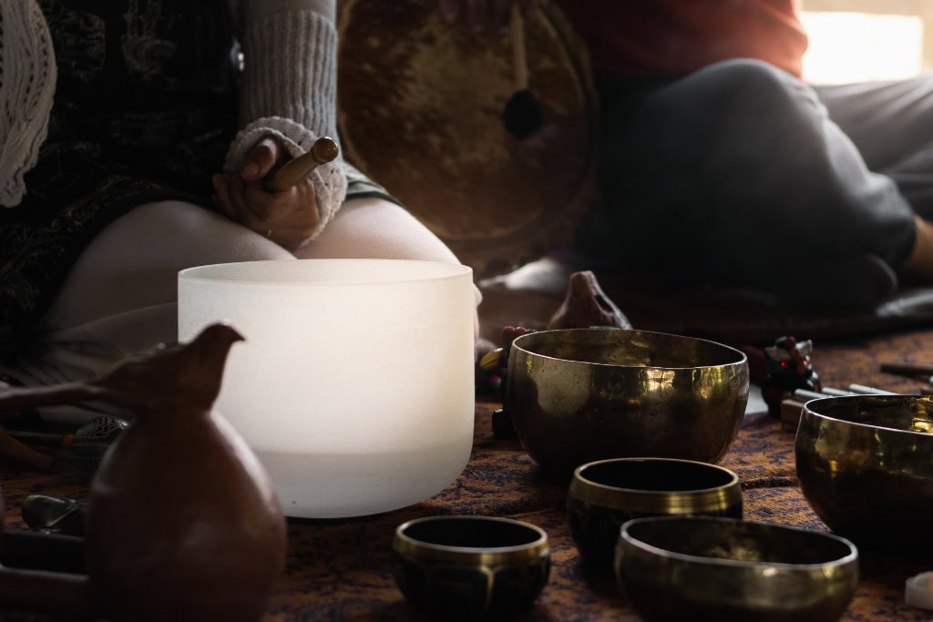 Cleansing Your House from Negative Energy: Everything You Need to Know! This image captures a meditative scene with a person using a mallet to play a large white singing bowl, surrounded by smaller brass bowls and other ritual objects. The dim lighting creates a tranquil, sacred atmosphere, suggesting a sound healing session or spiritual practice. Another person is partially visible in the background, holding a drum, adding to the contemplative and ceremonial mood.
