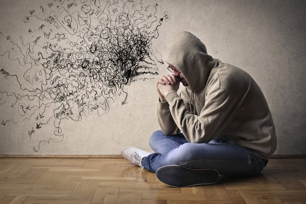 Cleansing Your House from Negative Energy: Everything You Need to Know! This image depicts a person wearing a hoodie, sitting on the floor with their head resting on their hands, appearing deep in thought or distress. On the wall next to them, chaotic scribbles and lines emerge from their head, symbolizing mental overwhelm or anxiety. The contrast between the person and the disorganized drawing emphasizes inner turmoil and confusion.