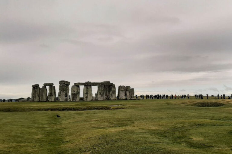 This image shows Stonehenge, the iconic prehistoric monument in England, with a cloudy sky overhead. The ancient stone circle is surrounded by a large grassy field, and a line of visitors can be seen in the distance walking around the monument. A single bird is visible in the foreground on the grass.