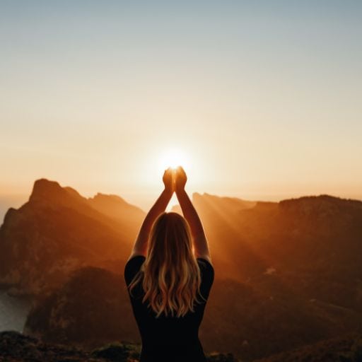 This image depicts a woman with long, wavy blonde hair standing with her back to the camera, raising her arms toward the sky as she frames the sun between her hands. The sun is setting or rising behind a range of rugged mountains, casting a golden glow across the landscape, creating a peaceful and contemplative scene. The woman's pose suggests a connection with nature, reflecting a sense of mindfulness or gratitude.