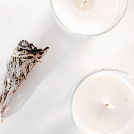 Cleansing Your House from Negative Energy: Everything You Need to Know! A bundle of sage and two white candles placed on a clean white surface, creating a serene and minimalistic setup often used for home energy cleansing rituals.