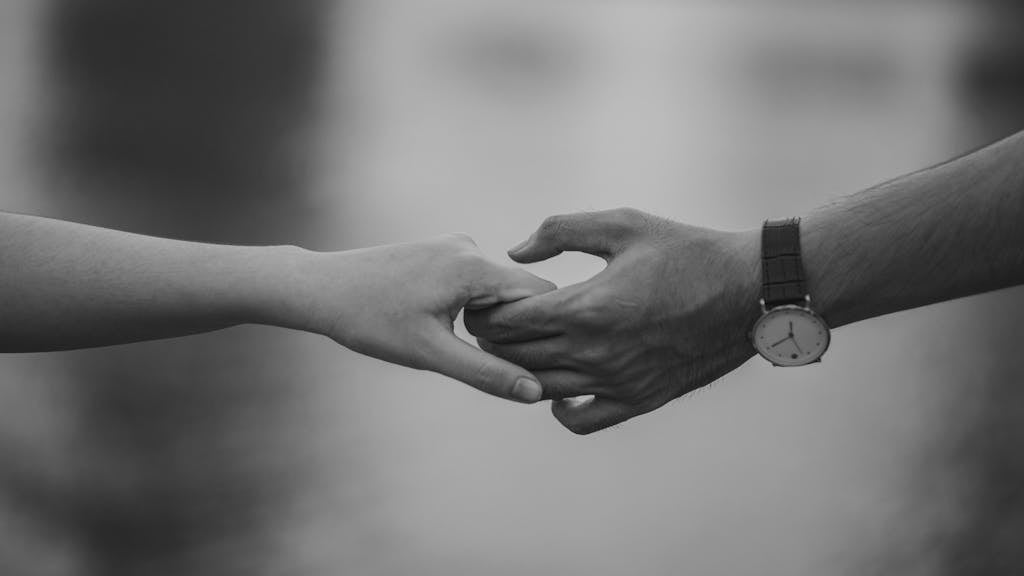 175 Angel Number - Meaning in Life Image of black-and-white photograph of a couple standing side by side, gently holding hands. Their fingers are intertwined, symbolizing connection and affection. The contrast of light and shadows highlights the tenderness of the moment, with their silhouettes softly blending into the background.