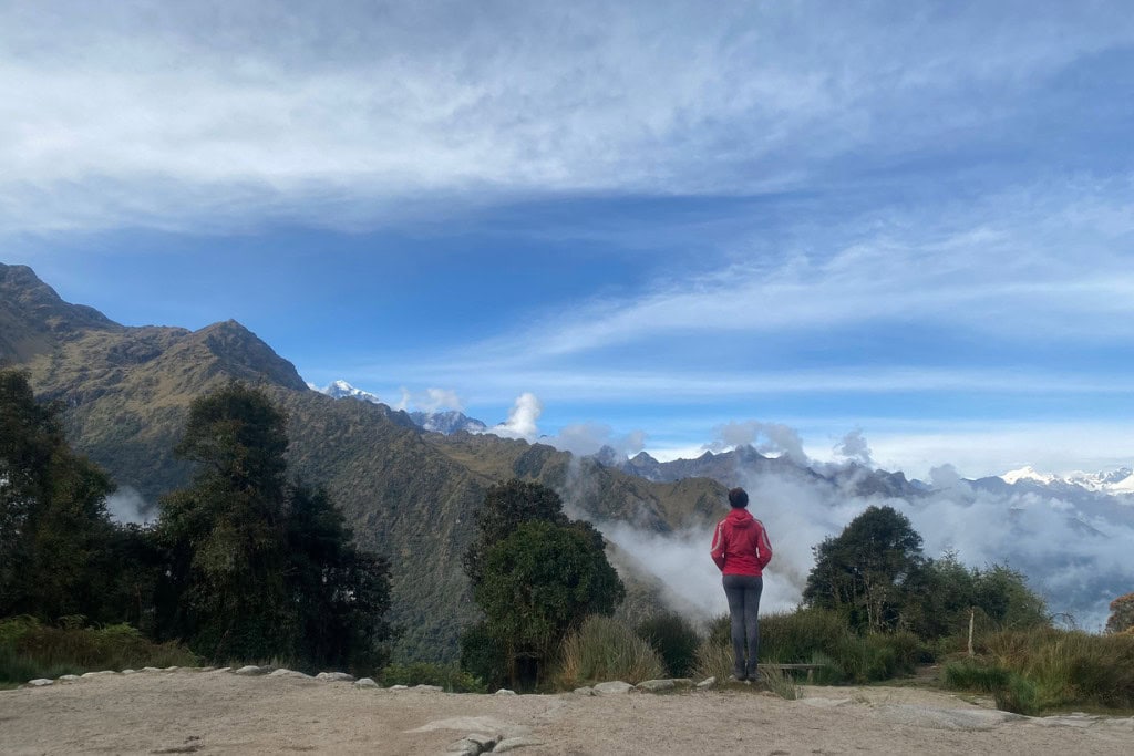 A person wearing a red jacket stands on a scenic overlook, facing the breathtaking mountain landscape and mist-shrouded valleys of Huancabamba in Peru. The sky above is expansive with streaks of white clouds, complementing the lush greenery and rugged peaks below.