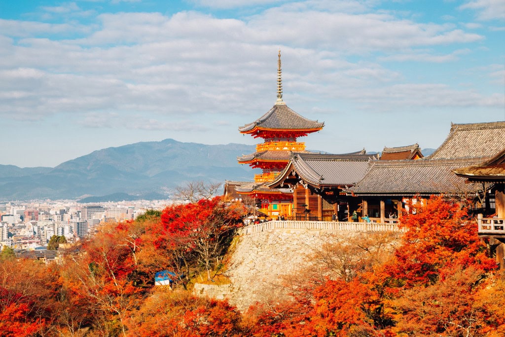 The image showcases Kiyomizu-dera temple in Kyoto, Japan, during autumn, with vibrant red and orange foliage surrounding the historic wooden structure. The temple's elevated position provides a panoramic view of Kyoto’s cityscape and distant mountains, enhancing the seasonal beauty and cultural charm of this iconic site. This scene captures the essence of Kyoto's historical sites amidst fall colors.