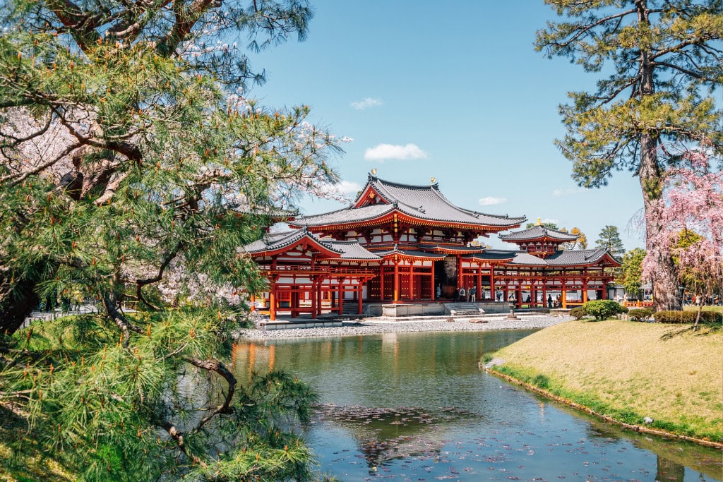 The image features the iconic Byodo-in Temple in Uji, Kyoto, Japan, reflected in a tranquil pond under a bright blue sky. Surrounded by lush greenery and cherry blossoms, the temple’s distinct red and white architecture stands out beautifully, capturing the elegance and historical significance of this UNESCO World Heritage site. This serene scene highlights the harmony of nature and traditional design typical of Kyoto’s temples.