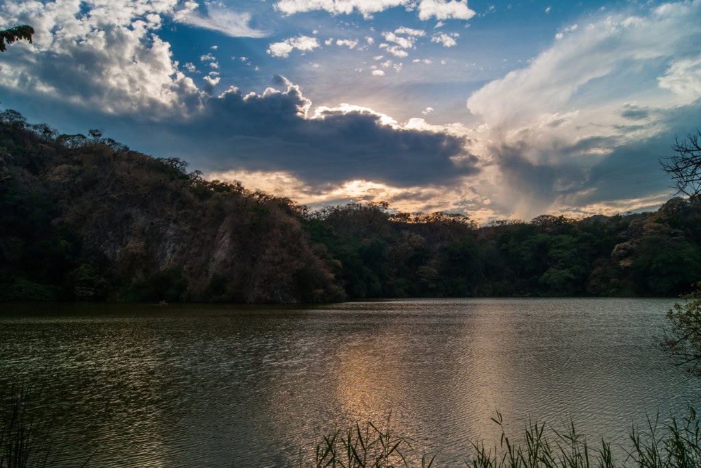 Huancabamba in Peru: Embark On A Spiritual Journey A serene lake surrounded by lush and rugged hills, reflecting the golden hues of the sky during sunset in Huancabamba, Peru. The dramatic clouds add depth to the sky, with streaks of light breaking through, creating a picturesque and tranquil landscape.