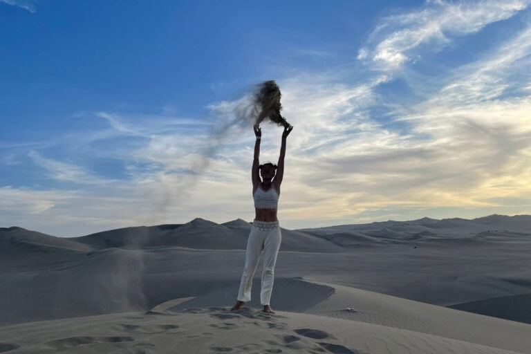 A person stands on a sand dune at dusk, raising their arms as they release sand into the air, symbolizing freedom and transformation. The expansive desert and vibrant sky set the stage for a powerful spiritual awakening journey, highlighting a moment of connection and self-discovery.