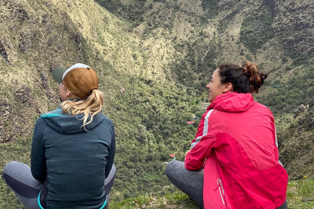 Huancabamba in Peru: Embark On A Spiritual Journey Two women sit on a grassy ledge overlooking a vast, misty mountain valley in Huancabamba, Peru. One wears a pink jacket and the other a green hoodie with a cap, both enjoying the breathtaking view and peaceful ambiance of the rugged landscape below.